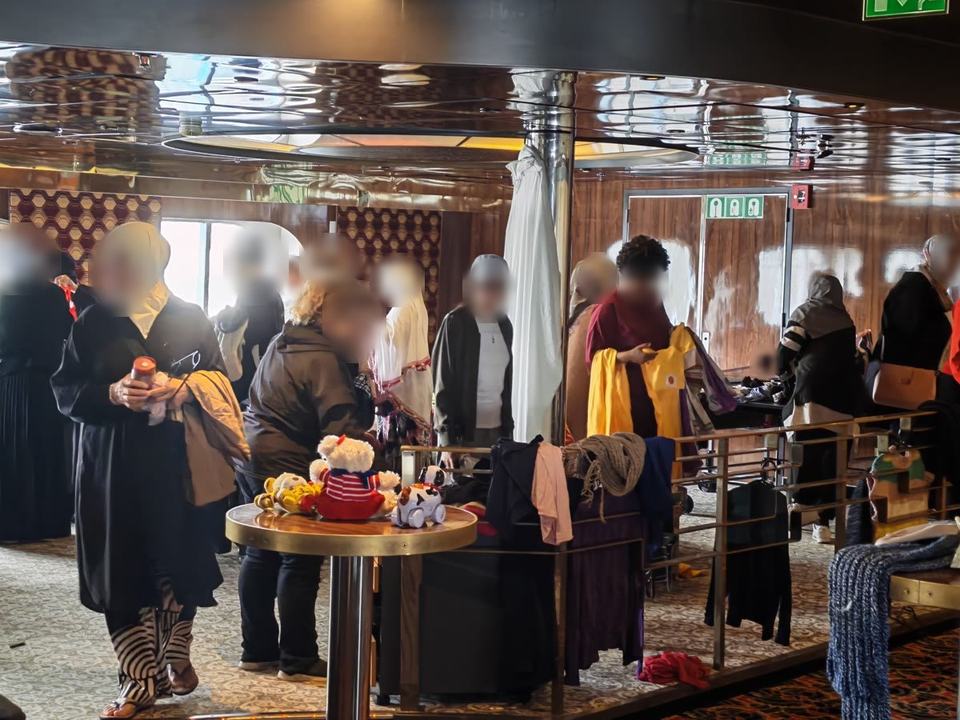 Women browsing clothes and toys on round tables and rails in a lounge on board a ship. Faces are blurred.