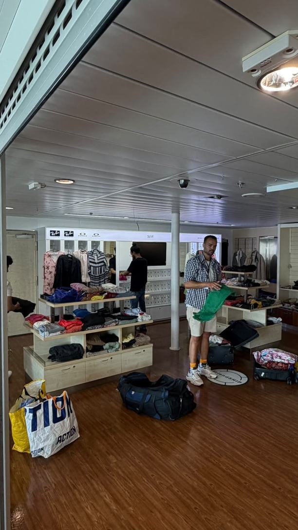 A shop space on a ship with shelves and rails of clothes, bags on the wooden floor, and our co-founder and a volunteer setting out clothes.