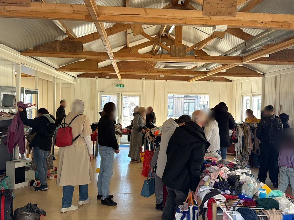 People browsing tables and shelves of clothes, shoes and toys in a large hall with a wooden roof.